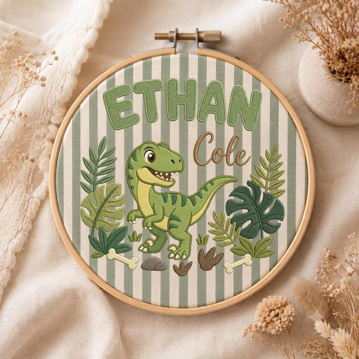 May include: A round wooden embroidery hoop containing fabric with vertical sage green and white stripes. The embroidery features a green dinosaur, tropical leaves, bones, and the text "ETHAN" in green block letters and "Cole" in brown script.