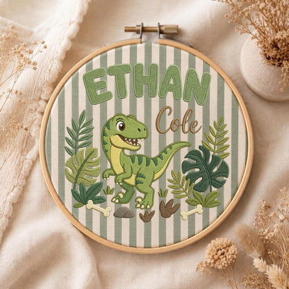 May include: A round wooden embroidery hoop containing fabric with vertical sage green and white stripes. The embroidery features a green dinosaur, tropical leaves, bones, and the text "ETHAN" in green block letters and "Cole" in brown script.