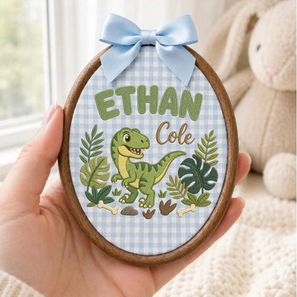 May include: An oval wooden embroidery hoop featuring a light blue and white gingham fabric background. The design includes the embroidered green text "ETHAN" and brown script "Cole" above a green dinosaur, tropical leaves, and small bones.