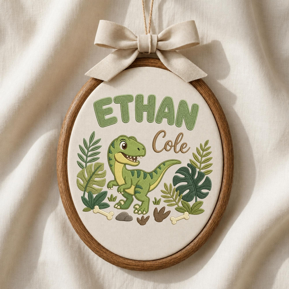 May include: An oval embroidery hoop wall hanging featuring a green dinosaur, tropical leaves, and bones stitched on cream fabric. The text reads ETHAN in green block letters and Cole in brown script. A beige fabric bow is attached to the top.