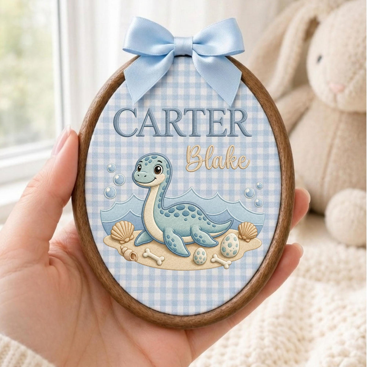 May include: An oval wooden embroidery hoop frame containing fabric with a light blue gingham pattern. The fabric features a cartoon blue dinosaur, seashells, and eggs, with the text CARTER and Blake embroidered in blue and gold script.