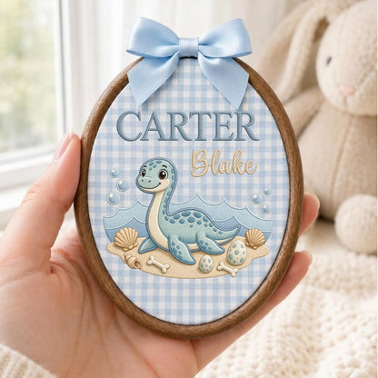 May include: An oval wooden embroidery hoop frame containing fabric with a light blue gingham pattern. The fabric features a cartoon blue dinosaur, seashells, and eggs, with the text CARTER and Blake embroidered in blue and gold script.