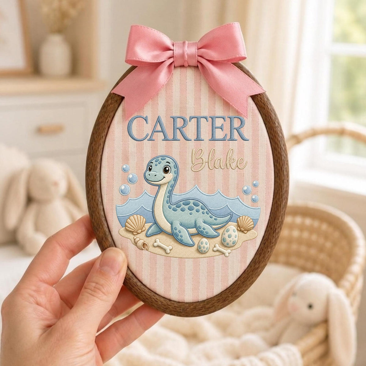 May include: An oval wooden embroidery hoop frame containing fabric with vertical pink and white stripes. The fabric features an embroidered blue dinosaur, seashells, and eggs, with the text CARTER Blake in blue and script lettering, topped with a pink bow.