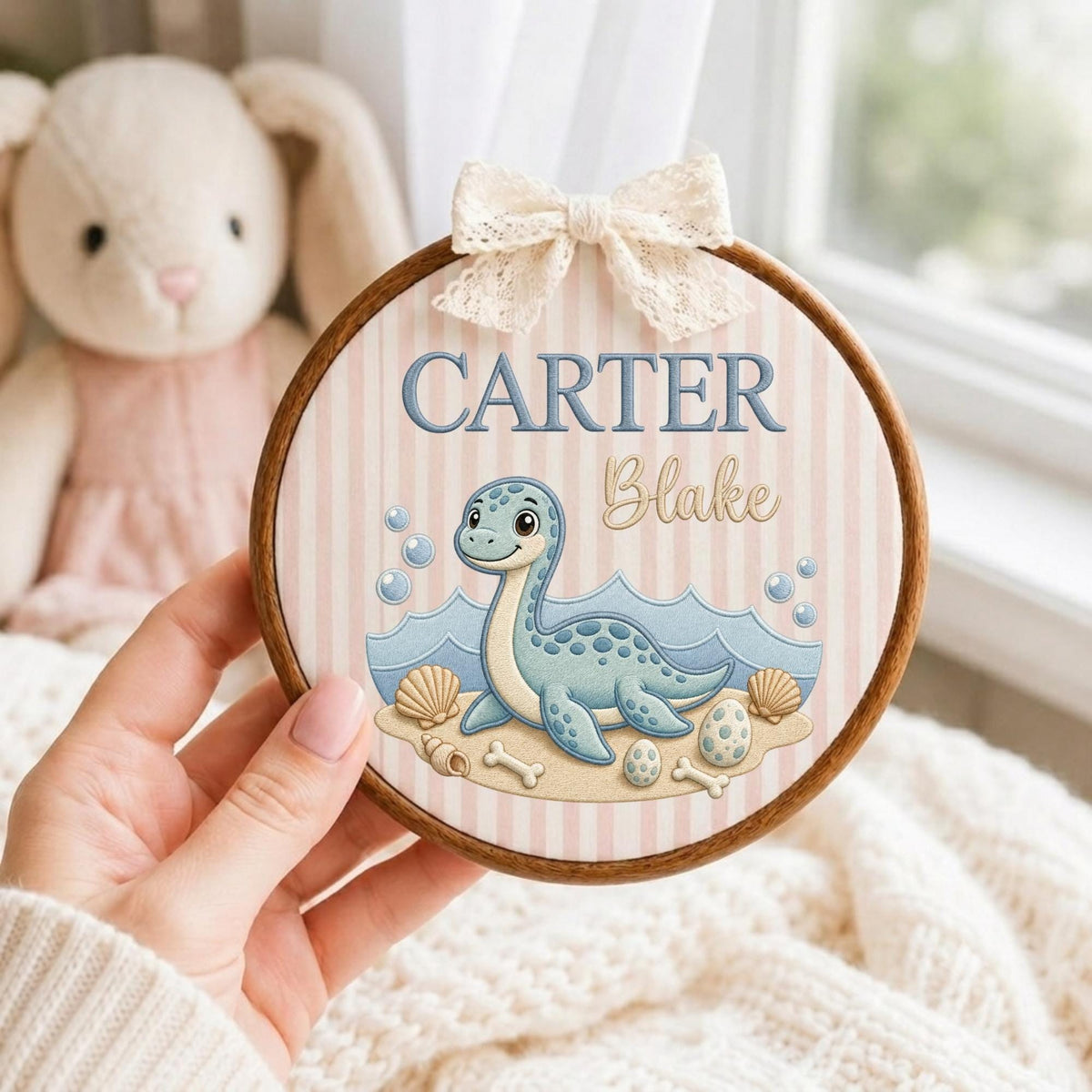 May include: A circular wooden embroidery hoop featuring a light pink and white striped fabric background. The design includes a blue dinosaur, seashells, and eggs on a sandy beach with the text CARTER Blake in blue and cursive script, topped with a lace bow.