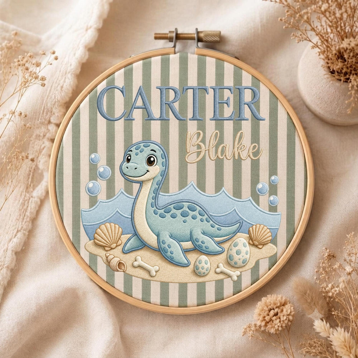 May include: A round wooden embroidery hoop containing fabric with vertical sage green and white stripes. The design features a blue cartoon dinosaur, seashells, and eggs on a sandy beach. The text reads CARTER in blue and Blake in script.