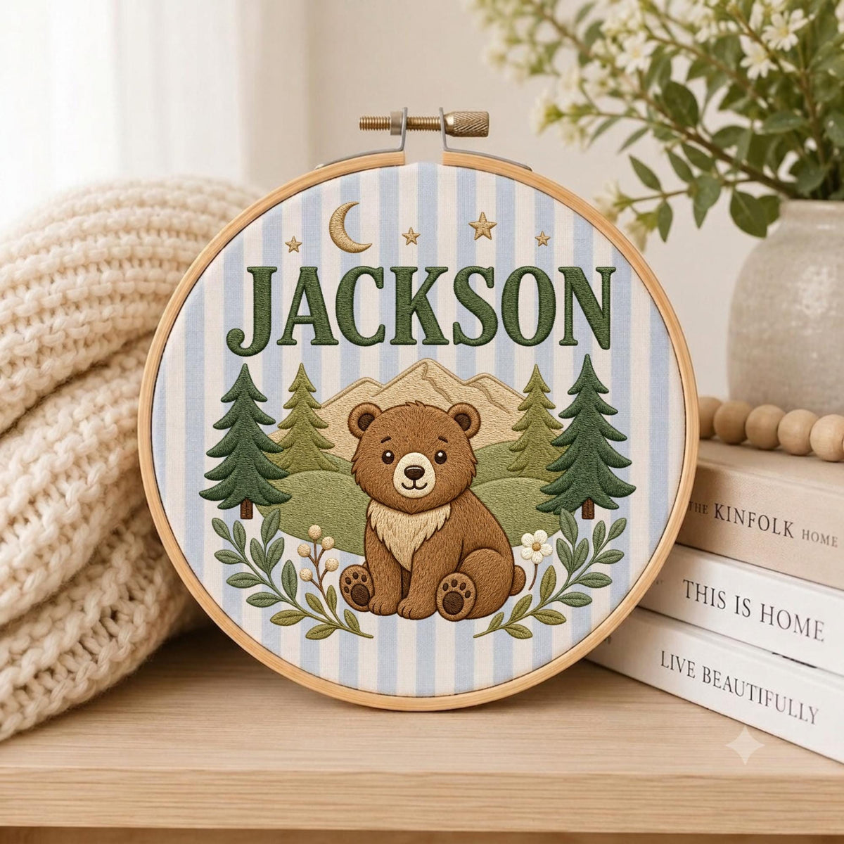 May include: A circular embroidery hoop featuring a light blue and white striped fabric background. The design includes a brown bear sitting in a forest with green pine trees, mountains, a crescent moon, stars, and the name JACKSON in green.