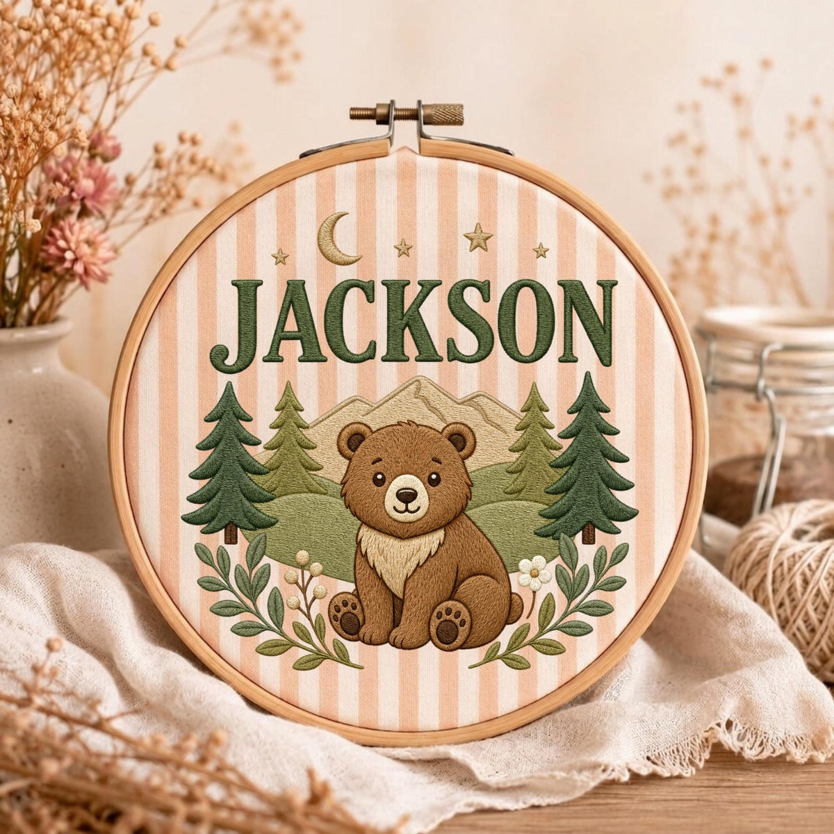 May include: A circular wooden embroidery hoop containing fabric with vertical light peach stripes. The embroidery features a brown bear sitting in a forest with green pine trees, mountains, a crescent moon, stars, and the name JACKSON in green.