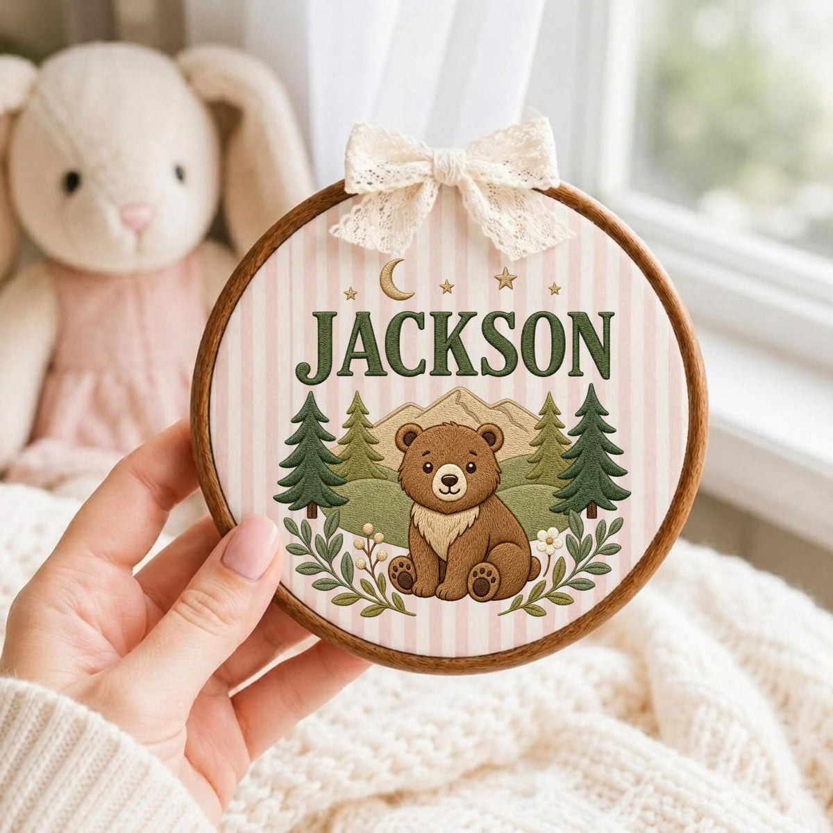 May include: A circular wooden embroidery hoop frame containing fabric with a light pink and white vertical striped pattern. The design features a brown bear, green pine trees, mountains, a crescent moon, stars, and the name JACKSON in green text.