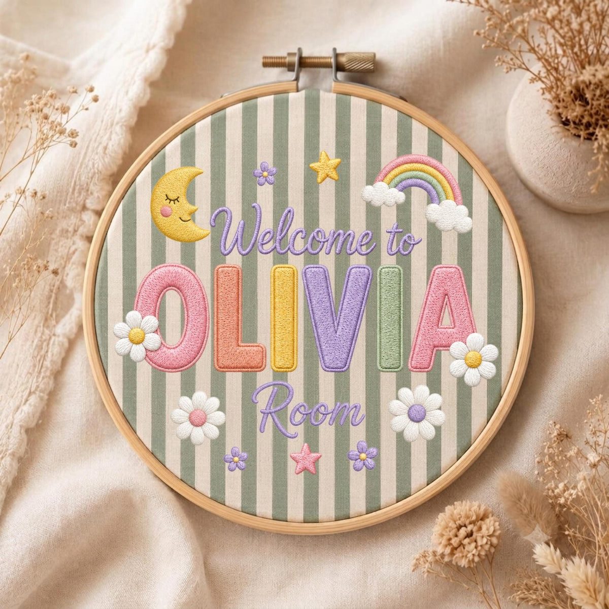May include: A circular wooden embroidery hoop containing fabric with green and white vertical stripes. The embroidery features the text Welcome to OLIVIA Room in purple, with a yellow crescent moon, a rainbow, and white flowers.