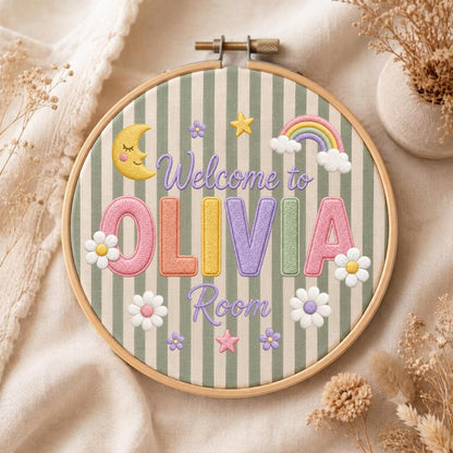 May include: A circular wooden embroidery hoop containing fabric with green and white vertical stripes. The embroidery features the text Welcome to OLIVIA Room in purple, with a yellow crescent moon, a rainbow, and white flowers.