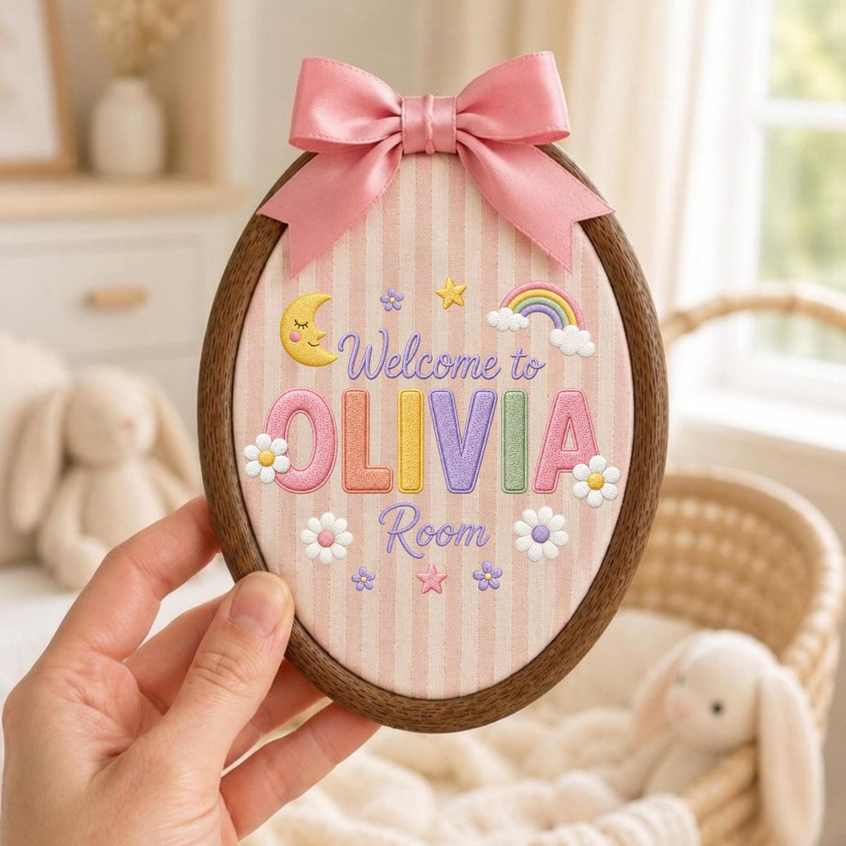 May include: An oval wooden embroidery hoop containing fabric with pink vertical stripes. The text "Welcome to OLIVIA Room" is embroidered in the center, surrounded by a yellow crescent moon, a rainbow, and white flowers, topped with a pink satin bow.