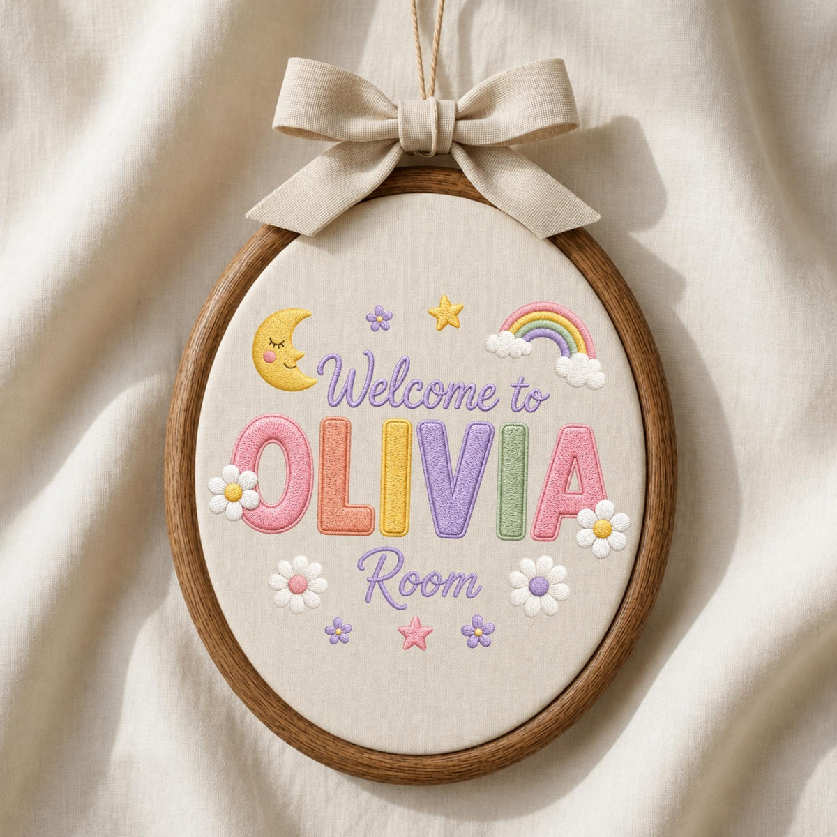 May include: An oval embroidery hoop wall hanging featuring the embroidered text Welcome to OLIVIA Room. The design includes a yellow crescent moon, a rainbow, stars, and white flowers on cream fabric, finished with a beige fabric bow at the top.