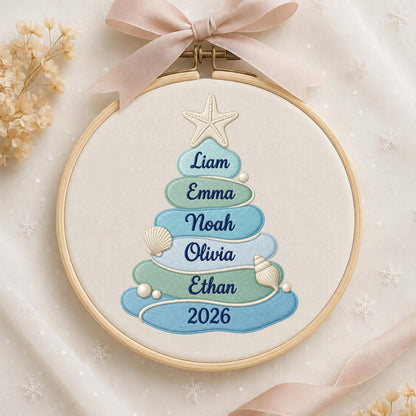 May include: A circular wooden embroidery hoop containing a fabric panel with a stitched Christmas tree design. The tree is composed of stacked blue and green oval shapes featuring the names Liam, Emma, Noah, Olivia, Ethan, and the year 2026.