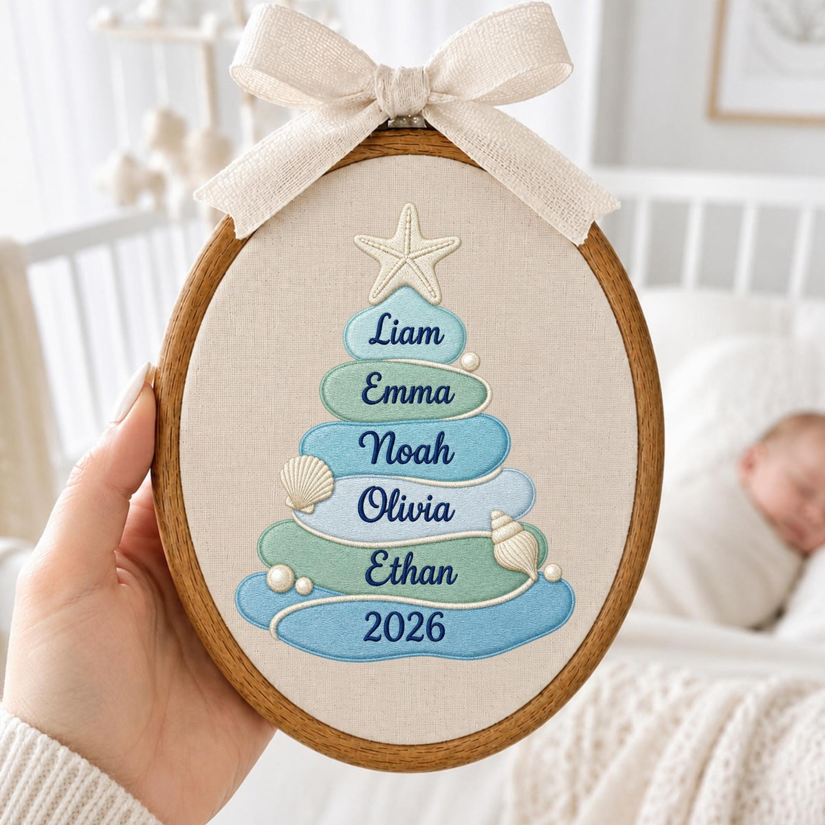 May include: An oval wooden embroidery hoop containing a beige fabric panel with a stylized Christmas tree design. The tree is composed of stacked blue and green fabric layers featuring the names Liam, Emma, Noah, Olivia, Ethan, and the year 2026.