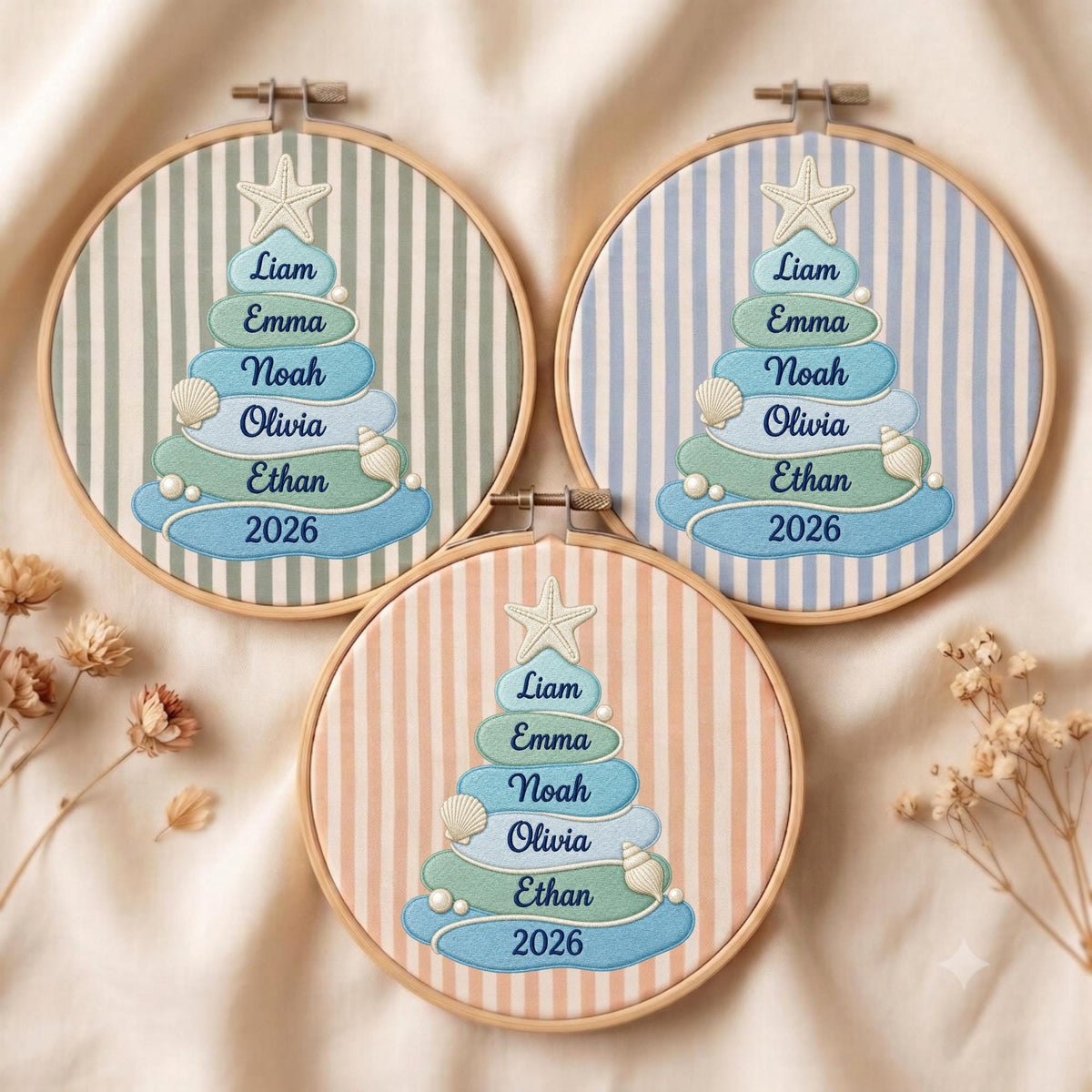 May include: Three circular wooden embroidery hoops featuring fabric with vertical stripes in sage green, light blue, and peach. Each hoop displays an embroidered Christmas tree design composed of layered blue and green shapes with the names Liam, Emma, Noah, Olivia, Ethan, and the year 2026, topped with a starfish and accented with seashells.