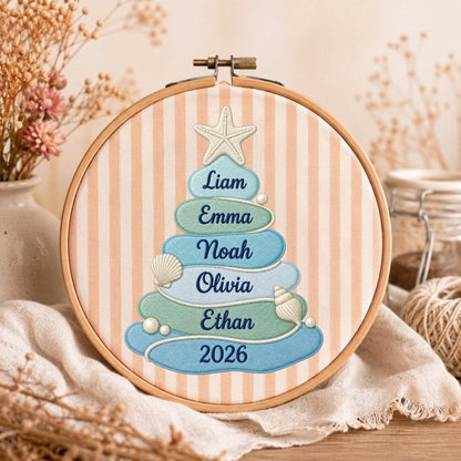 May include: A circular wooden embroidery hoop containing fabric with vertical peach and white stripes. The fabric features an embroidered tree shape made of stacked blue and green layers, topped with a starfish, seashells, and the names Liam, Emma, Noah, Olivia, Ethan, and 2026.