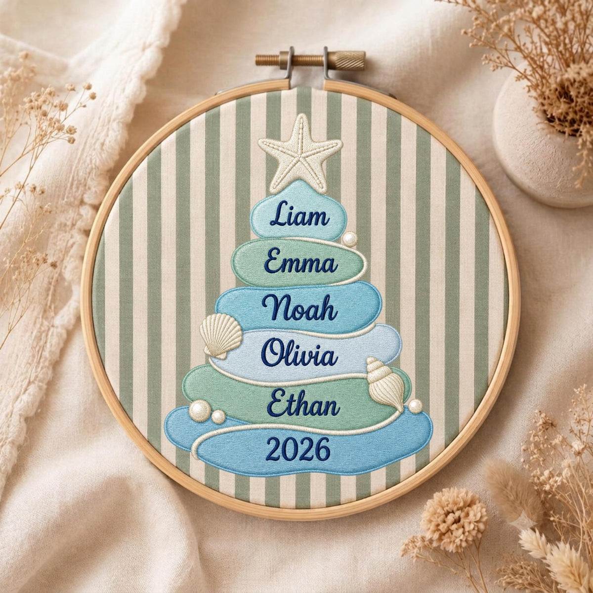 May include: A circular wooden embroidery hoop containing fabric with vertical sage green and white stripes. An embroidered Christmas tree shape is formed by stacked blue and green ovals featuring the names Liam, Emma, Noah, Olivia, Ethan, and 2026.