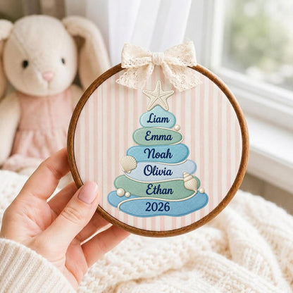 May include: A circular wooden embroidery hoop frame containing fabric with a vertical light pink striped pattern. A stylized Christmas tree made of blue and green rounded shapes features the names Liam, Emma, Noah, Olivia, Ethan and 2026.