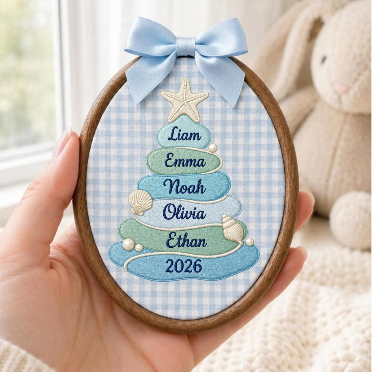 May include: An oval wooden embroidery hoop containing fabric with a light blue gingham pattern. The fabric features an embroidered tree shape made of blue and green layers, decorated with a starfish, seashells, pearls, and the names Liam, Emma, Noah, Olivia, Ethan, and 2026. A light blue satin ribbon bow is attached to the top.