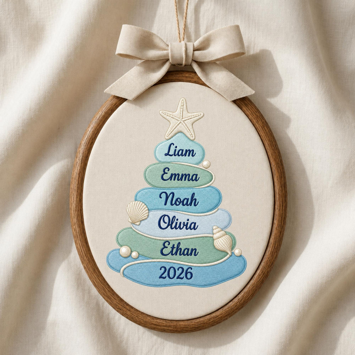 May include: An oval embroidery hoop containing a cream-colored fabric with a tree-shaped design made of blue and green layers. The tree features embroidered names Liam, Emma, Noah, Olivia, Ethan, and the year 2026, accented with a starfish and shells.