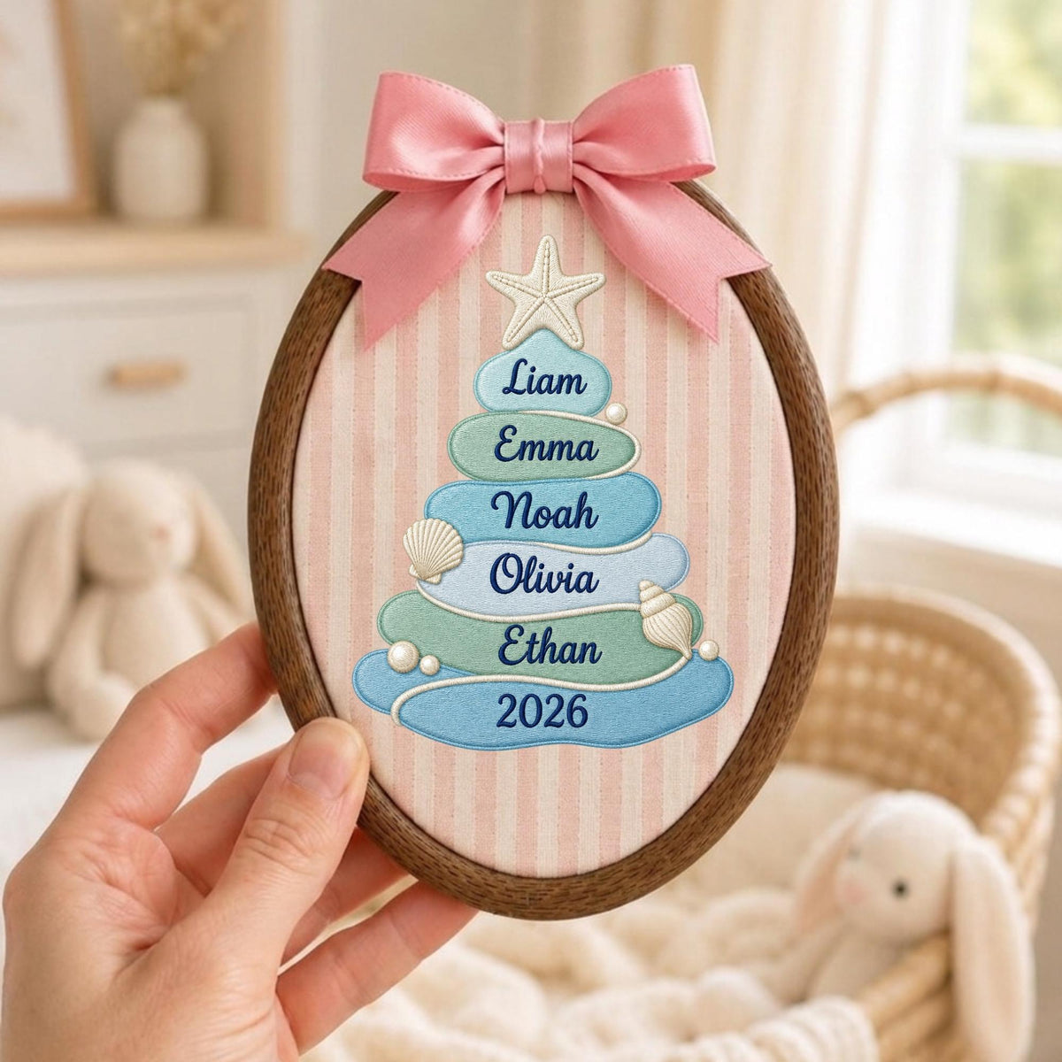 May include: An oval wooden embroidery hoop containing fabric with a pink and white striped pattern. A pink satin bow sits at the top. The fabric features an embroidered tree made of blue and green shapes with the names Liam, Emma, Noah, Olivia, Ethan, and the year 2026, accented by a starfish, seashells, and small white pearls.