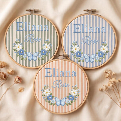 May include: Three circular wooden embroidery hoops featuring striped fabric in sage green, light blue, and peach. Each hoop displays the embroidered name Eliana Rose in blue script and block letters, accented by a floral wreath and a blue butterfly.