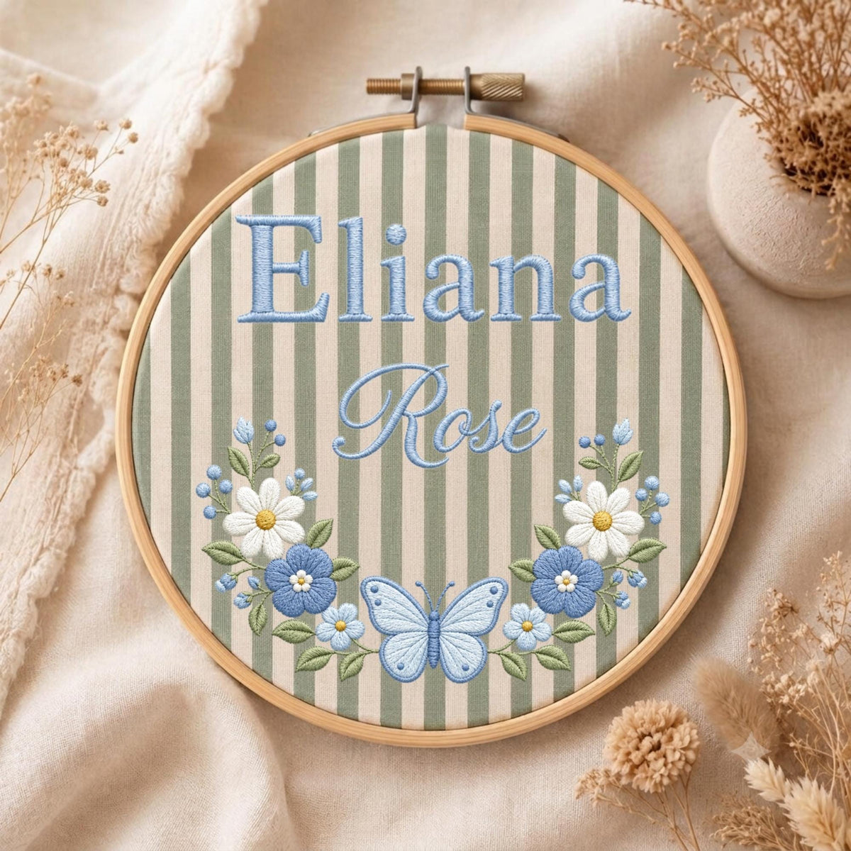 May include: A circular wooden embroidery hoop containing fabric with vertical sage green and cream stripes. The name Eliana Rose is embroidered in light blue thread. Below the text is a floral wreath with white and blue flowers and a butterfly.
