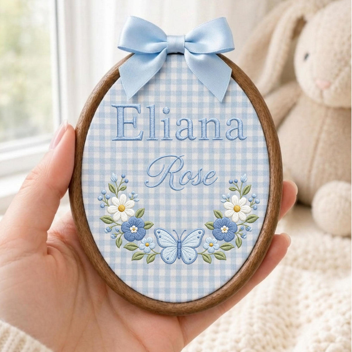 May include: An oval wooden embroidery hoop containing light blue and white gingham fabric. The fabric features the embroidered text Eliana Rose in blue, accented by a floral wreath with white and blue flowers and a central blue butterfly.