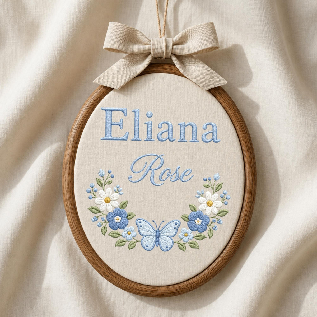 May include: An oval wooden embroidery hoop containing cream-colored fabric with the embroidered blue text Eliana Rose. Below the text is a floral wreath featuring white and blue flowers, green leaves, and a central light blue butterfly. A beige ribbon bow is attached to the top.