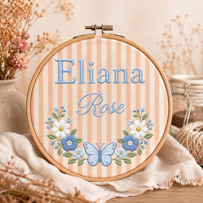 May include: A circular wooden embroidery hoop containing fabric with vertical peach and white stripes. The name Eliana Rose is embroidered in blue thread above a floral wreath featuring white and blue flowers and a central blue butterfly.