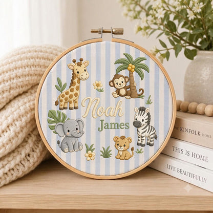 May include: A circular wooden embroidery hoop containing fabric with light blue and white vertical stripes. The fabric features embroidered illustrations of a giraffe, monkey, elephant, zebra, and lion, with the name Noah James in the center.