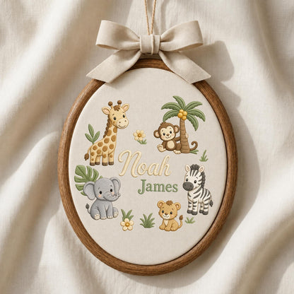May include: An oval wooden embroidery hoop featuring a cream fabric background with embroidered illustrations of a giraffe, monkey, elephant, zebra, and lion. The text "Noah James" is centered in the middle, and a beige ribbon bow is tied at the top.