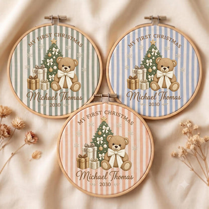 May include: Three circular wooden embroidery hoops featuring fabric with vertical stripes in sage green, light blue, and soft peach. Each hoop displays a printed illustration of a teddy bear, a decorated Christmas tree, gift boxes, and the text "MY FIRST CHRISTMAS", "Michael Thomas", and "2030".