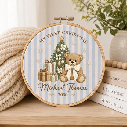 May include: A circular embroidery hoop featuring a light blue and white striped fabric background. The design includes a Christmas tree, a brown teddy bear with a white bow, gift boxes, and the text My First Christmas, Michael Thomas, 2030.