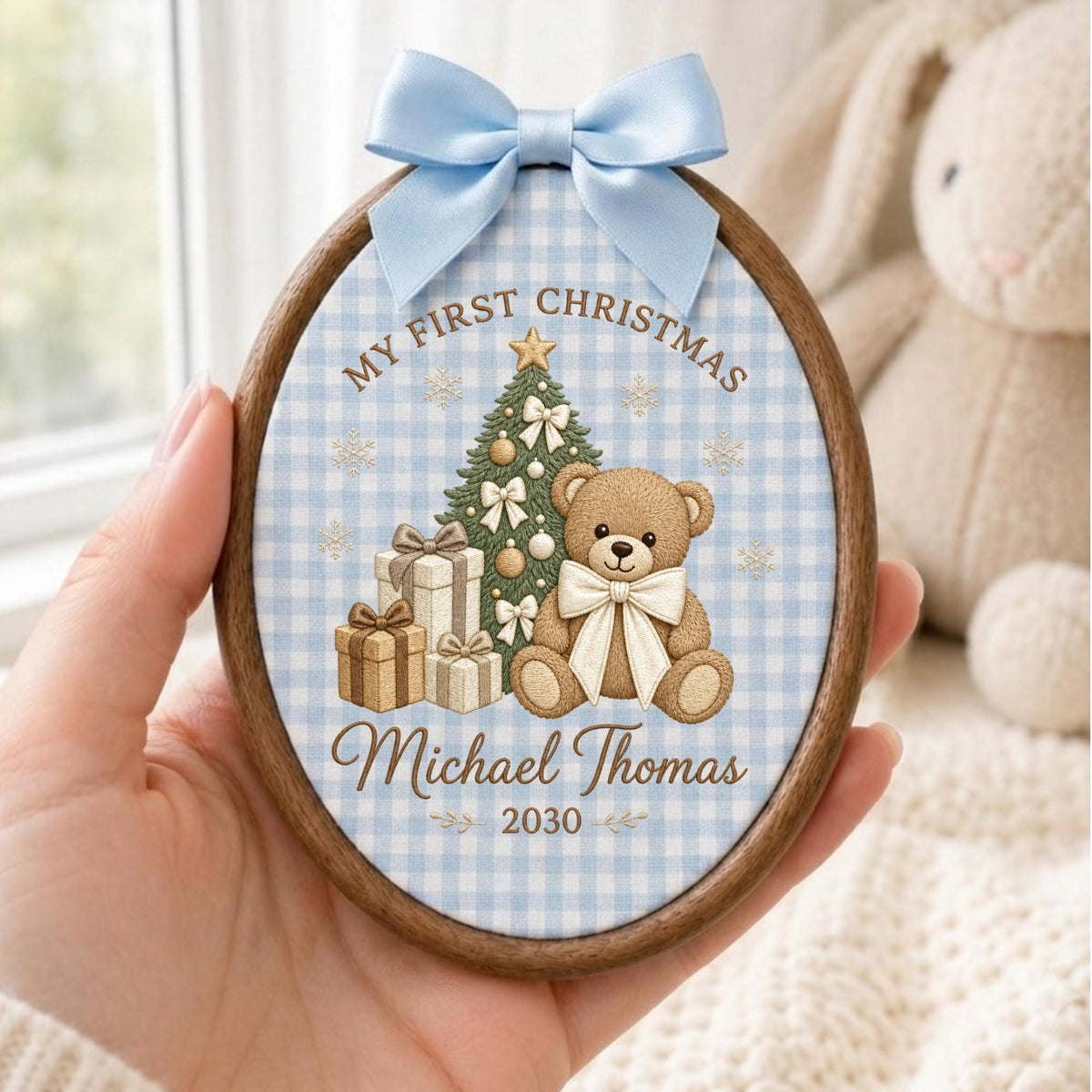 May include: An oval wooden ornament featuring a light blue gingham fabric background. The design includes a brown teddy bear, a decorated Christmas tree, and gift boxes. Text reads: MY FIRST CHRISTMAS, Michael Thomas, and 2030. A light blue satin bow is attached to the top.