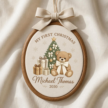 May include: An oval wooden embroidery hoop ornament featuring a beige fabric center with a brown teddy bear, a decorated Christmas tree, and gift boxes. The text reads My First Christmas, Michael Thomas, and 2030. A beige fabric bow is attached at the top.