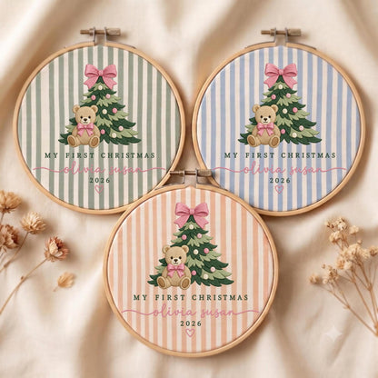 May include: Three circular wooden embroidery hoops featuring striped fabric in sage green, light blue, and peach. Each displays a Christmas tree with a pink bow, a teddy bear, and the text "MY FIRST CHRISTMAS", "olivia susan", and "2026".