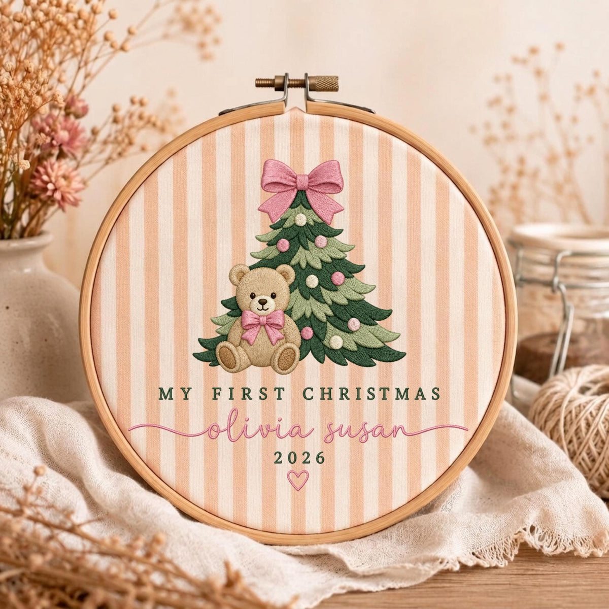 May include: A circular wooden embroidery hoop framing fabric with vertical peach and white stripes. The design features an embroidered green Christmas tree with pink ornaments, a teddy bear with a pink bow, and the text My First Christmas Olivia Susan 2026.
