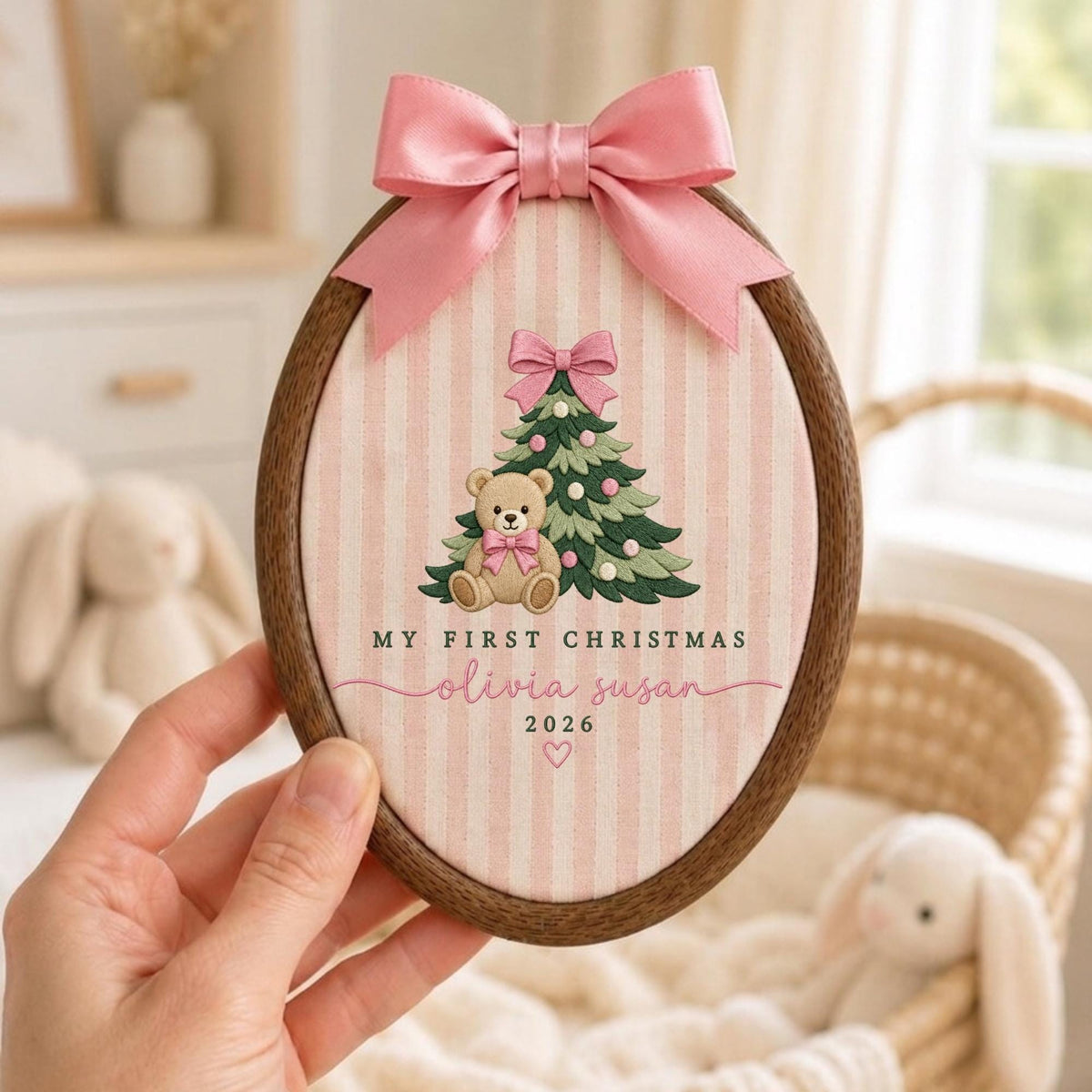 May include: An oval wooden embroidery hoop frame containing fabric with pink and white vertical stripes. The design features a teddy bear, a Christmas tree, and the text "MY FIRST CHRISTMAS olivia susan 2026" with a pink satin bow on top.