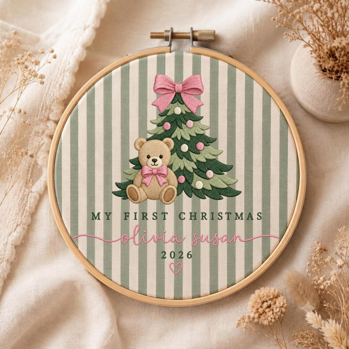 May include: A circular wooden embroidery hoop containing fabric with green and white vertical stripes. The embroidery features a green Christmas tree with a pink bow, a brown teddy bear with a pink bow, and the text MY FIRST CHRISTMAS, olivia susan, 2026, and a small heart.