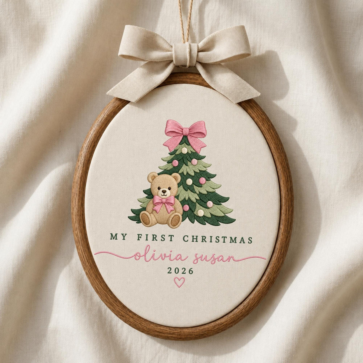 May include: An oval wooden embroidery hoop containing cream fabric with a stitched Christmas tree, a teddy bear with a pink bow, and the text MY FIRST CHRISTMAS, olivia susan, 2026, and a heart. A beige ribbon bow is tied at the top.