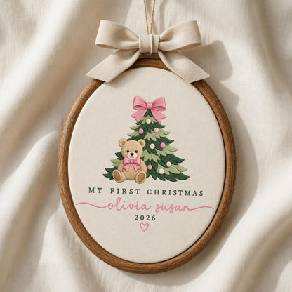 May include: An oval wooden embroidery hoop containing cream fabric with a stitched Christmas tree, a teddy bear with a pink bow, and the text MY FIRST CHRISTMAS, olivia susan, 2026, and a heart. A beige ribbon bow is tied at the top.