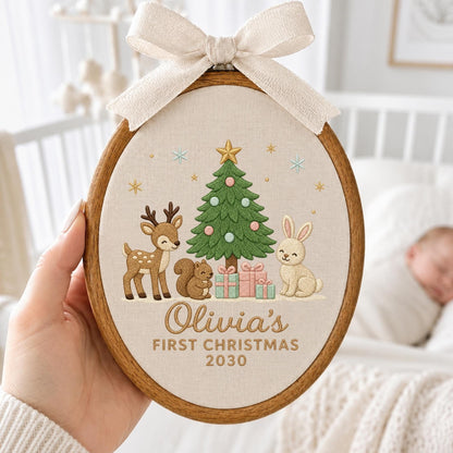 May include: An oval wooden embroidery hoop containing a beige fabric panel with a stitched Christmas tree, a deer, a squirrel, a rabbit, and gifts. The text reads Olivia's FIRST CHRISTMAS 2030. A cream-colored fabric bow is attached to the top of the hoop.