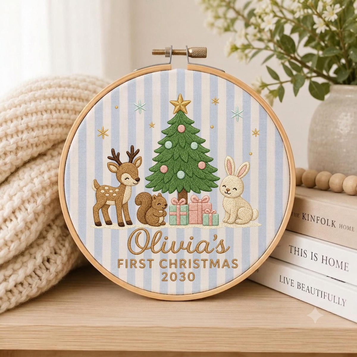 May include: A circular embroidery hoop featuring a light blue and white striped fabric background. The design includes a green Christmas tree, a brown deer, a brown squirrel, and a beige rabbit. Text reads: Olivia's FIRST CHRISTMAS 2030.
