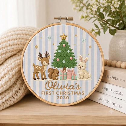 May include: A circular embroidery hoop featuring a light blue and white striped fabric background. The design includes a green Christmas tree, a brown deer, a brown squirrel, and a beige rabbit. Text reads: Olivia's FIRST CHRISTMAS 2030.