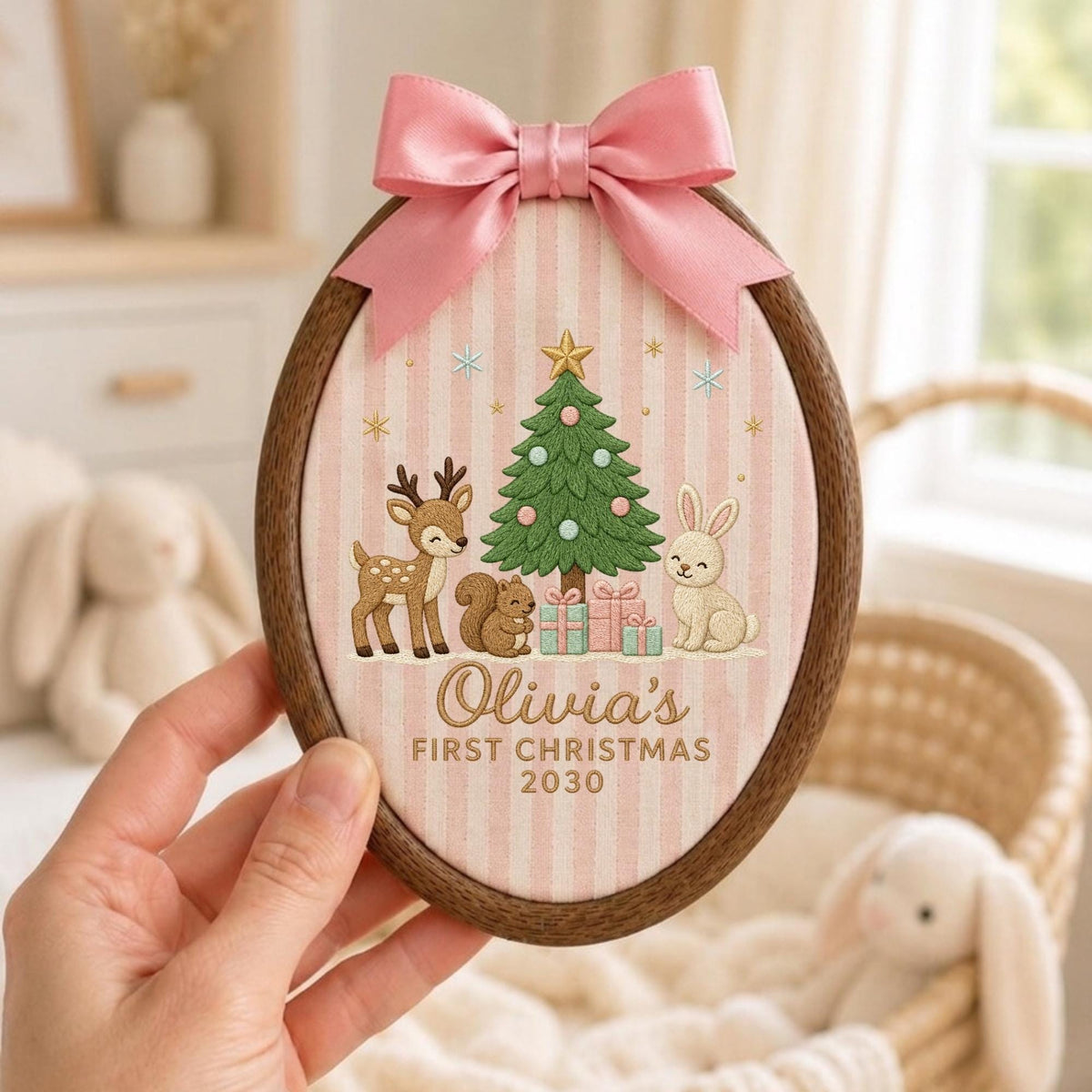 May include: An oval wooden embroidery hoop frame containing fabric with a pink and white striped pattern. The design features an embroidered Christmas tree, a deer, a squirrel, a rabbit, and gifts. Text reads: Olivia's FIRST CHRISTMAS 2030. A pink satin bow is attached to the top.