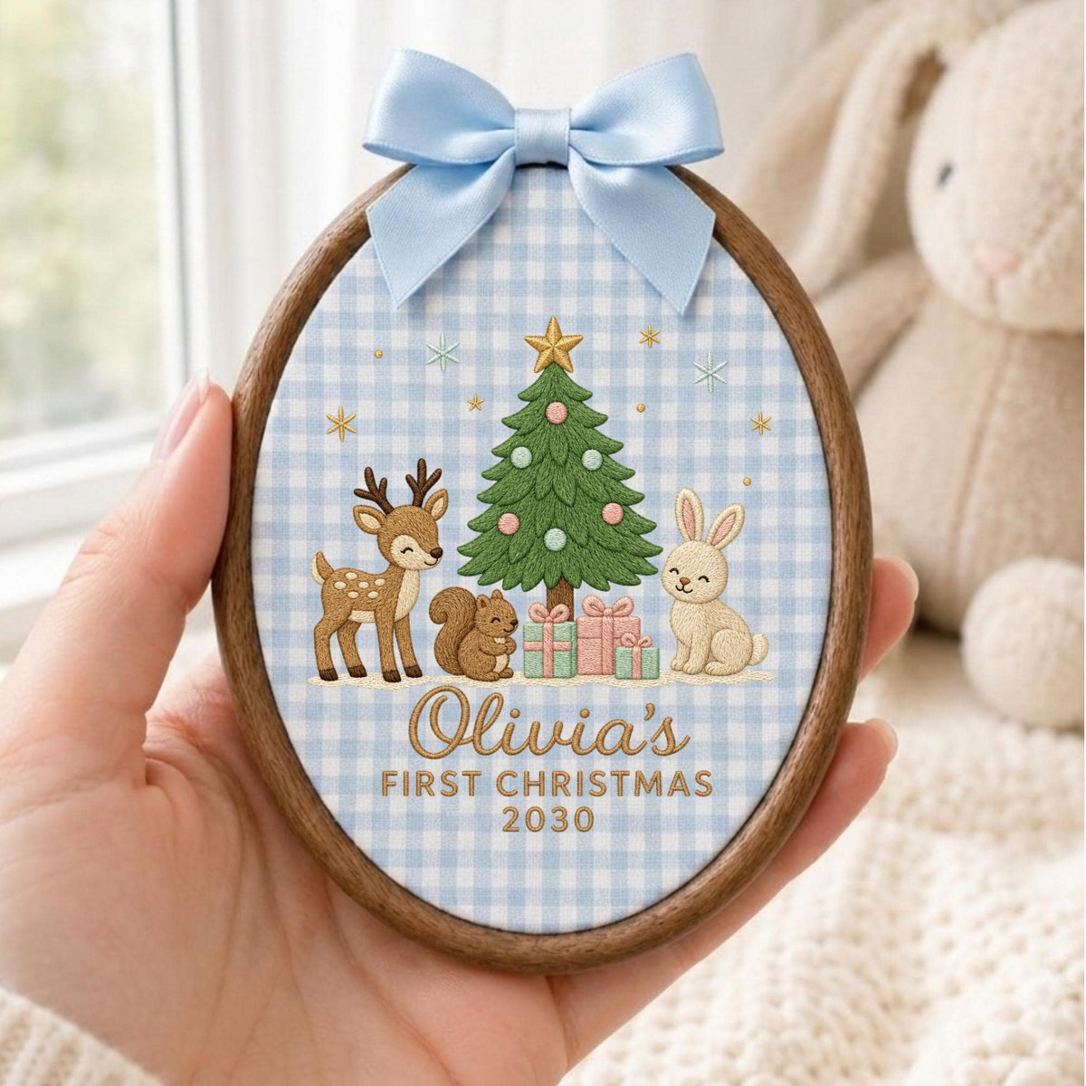 May include: An oval wooden embroidery hoop ornament featuring a light blue gingham fabric background. The design includes an embroidered green Christmas tree, a deer, a squirrel, a rabbit, and gifts. Text reads: Olivia's FIRST CHRISTMAS 2030.