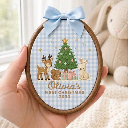May include: An oval wooden embroidery hoop ornament featuring a light blue gingham fabric background. The design includes an embroidered green Christmas tree, a deer, a squirrel, a rabbit, and gifts. Text reads: Olivia's FIRST CHRISTMAS 2030.