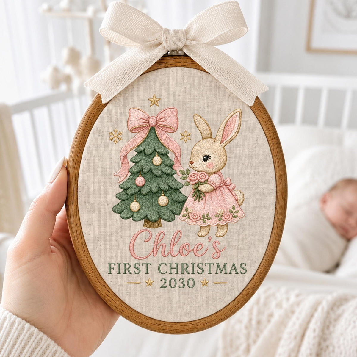 May include: An oval wooden embroidery hoop featuring a beige fabric background with a green Christmas tree, a pink bow, and a bunny in a pink dress. The text reads Chloe's FIRST CHRISTMAS 2030. A cream-colored ribbon bow is tied at the top.