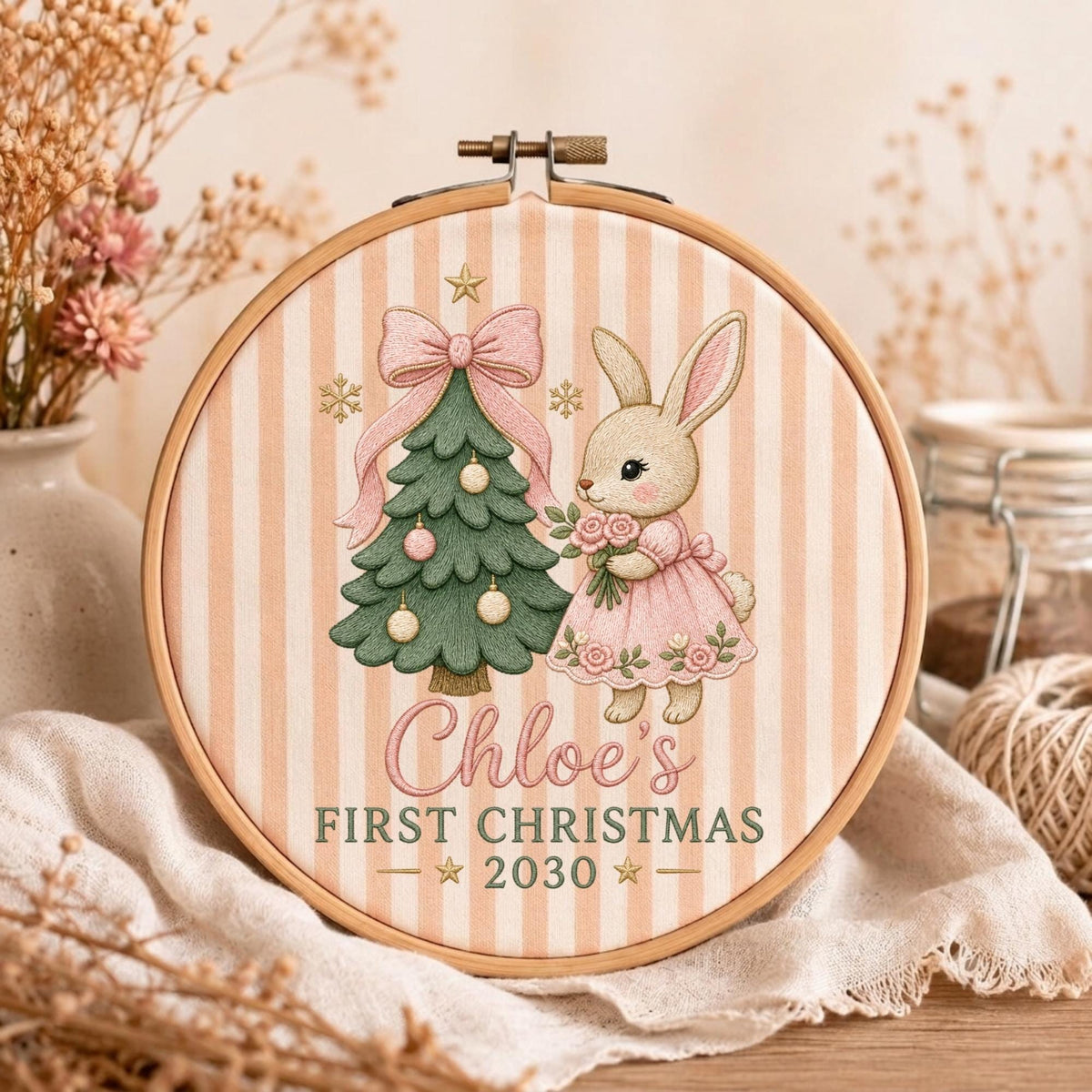 May include: A round wooden embroidery hoop containing fabric with a beige and white vertical striped pattern. The design features a green Christmas tree with a pink bow, a rabbit in a pink dress holding flowers, and the text Chloe's FIRST CHRISTMAS 2030.
